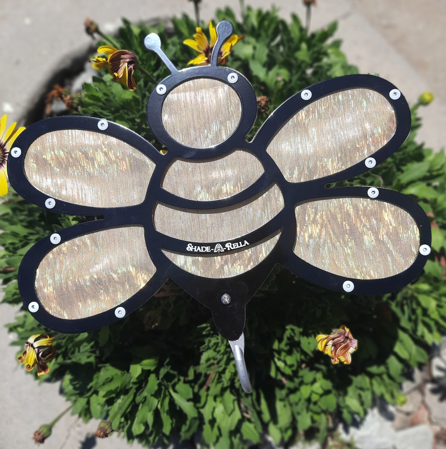 The Shade-A-Rella 10 inch small Gold Shimmer Bee, a bee-shaped garden ornament with translucent wings and black frame, adds a charming accent beside the 100% plant shade on a sunny day among green foliage and yellow flowers.