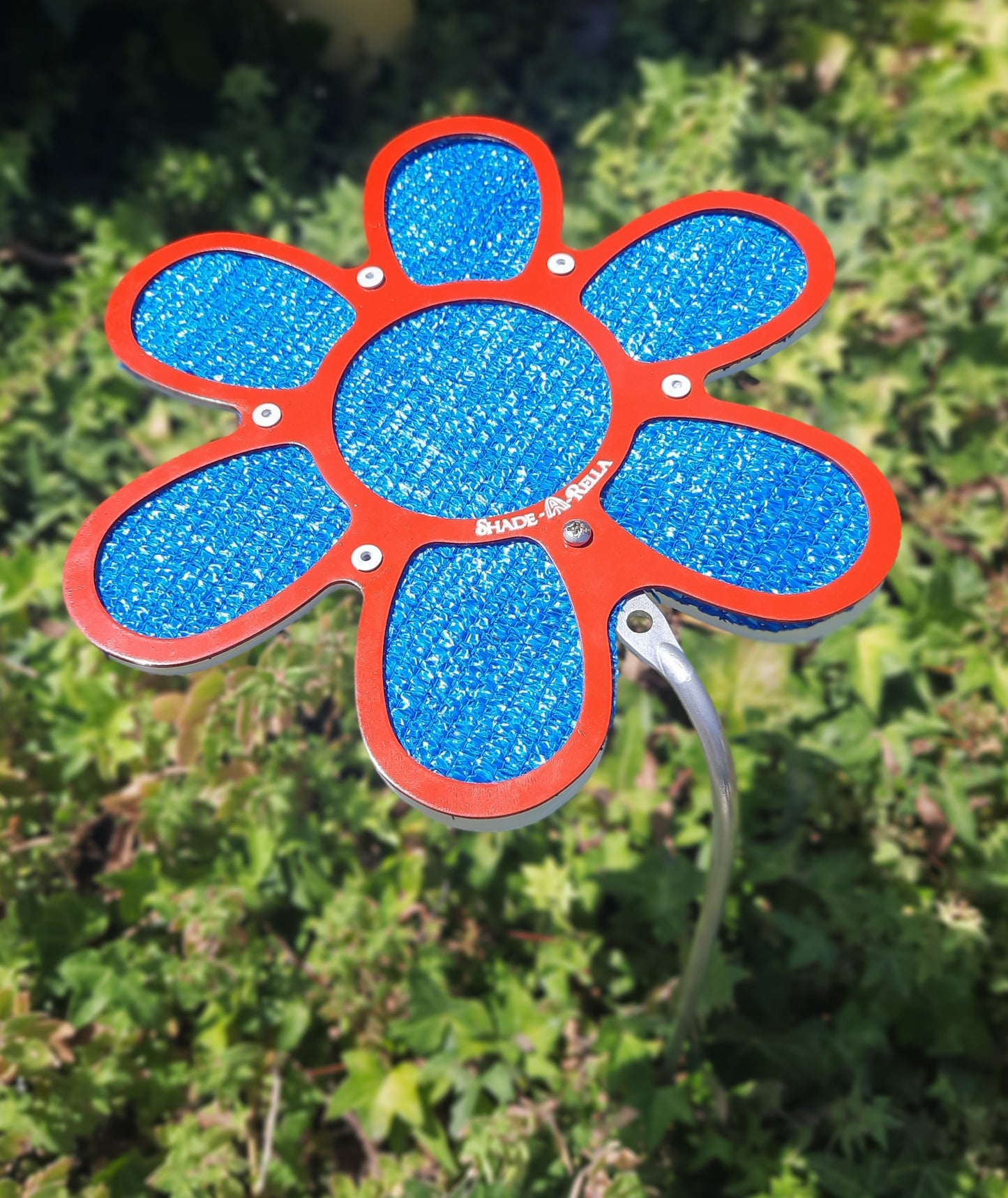 The Shade-A-Rella 7.5 inch small Flower features a red frame with blue 86% shade cloth petals, providing stylish shade above green foliage.