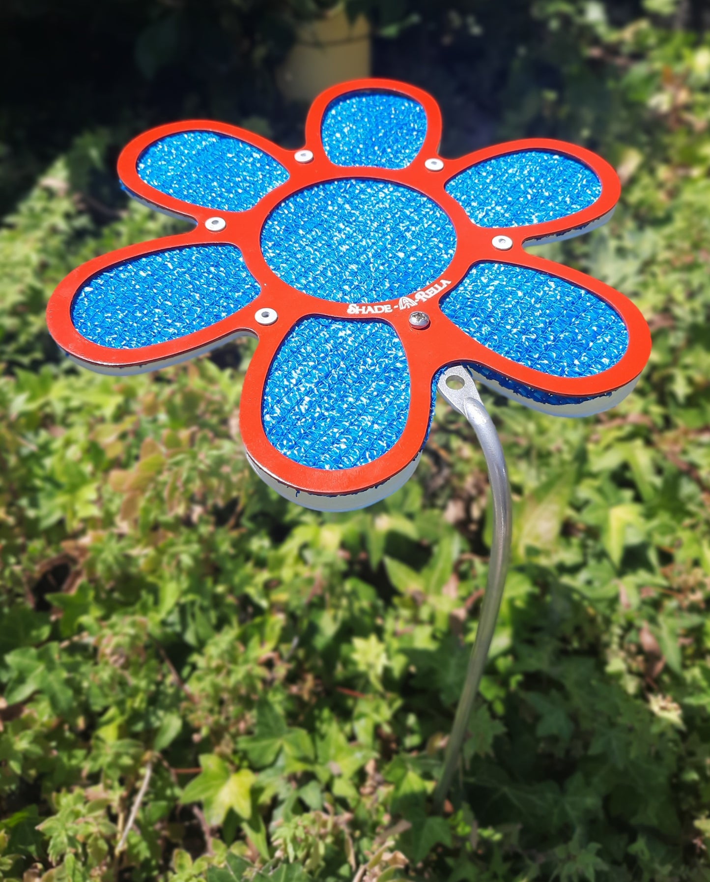 The Shade-A-Rella 7.5 inch small Flower features a red frame with blue 86% shade cloth, creating a charming metal ornament that stands among greenery to offer gentle shade and vibrant decorative appeal.