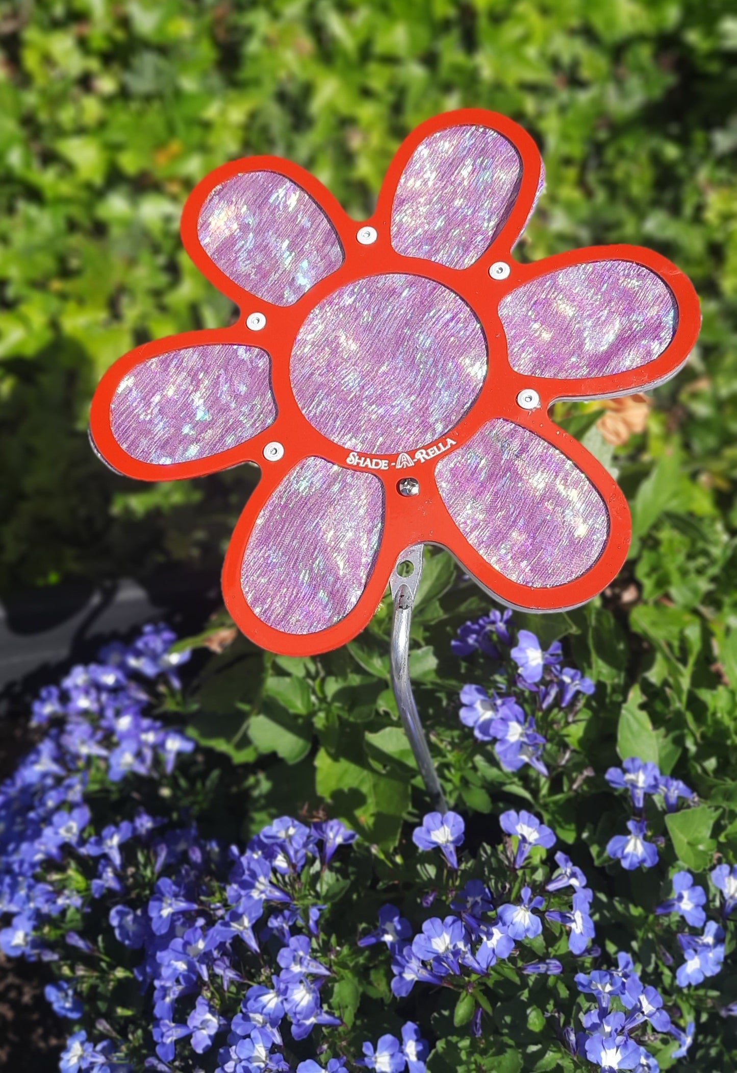 A 7.5 small Shimmer Flowers pinwheel from Shade-A-Rella stands among blooming purple and white shade plants, adding a touch of red and pink metallic shine with green foliage in the background.