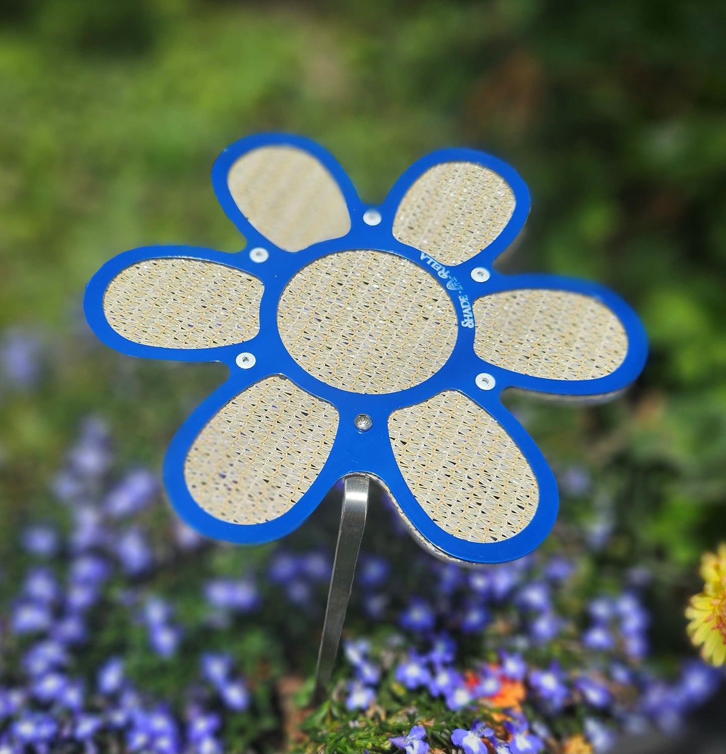 A 7.5-inch blue flower-shaped plant shade by Shade-A-Rella, made with yellow/tan 86% shade cloth, is mounted on a metal stake in a garden surrounded by green plants, window boxes, and small purple flowers.