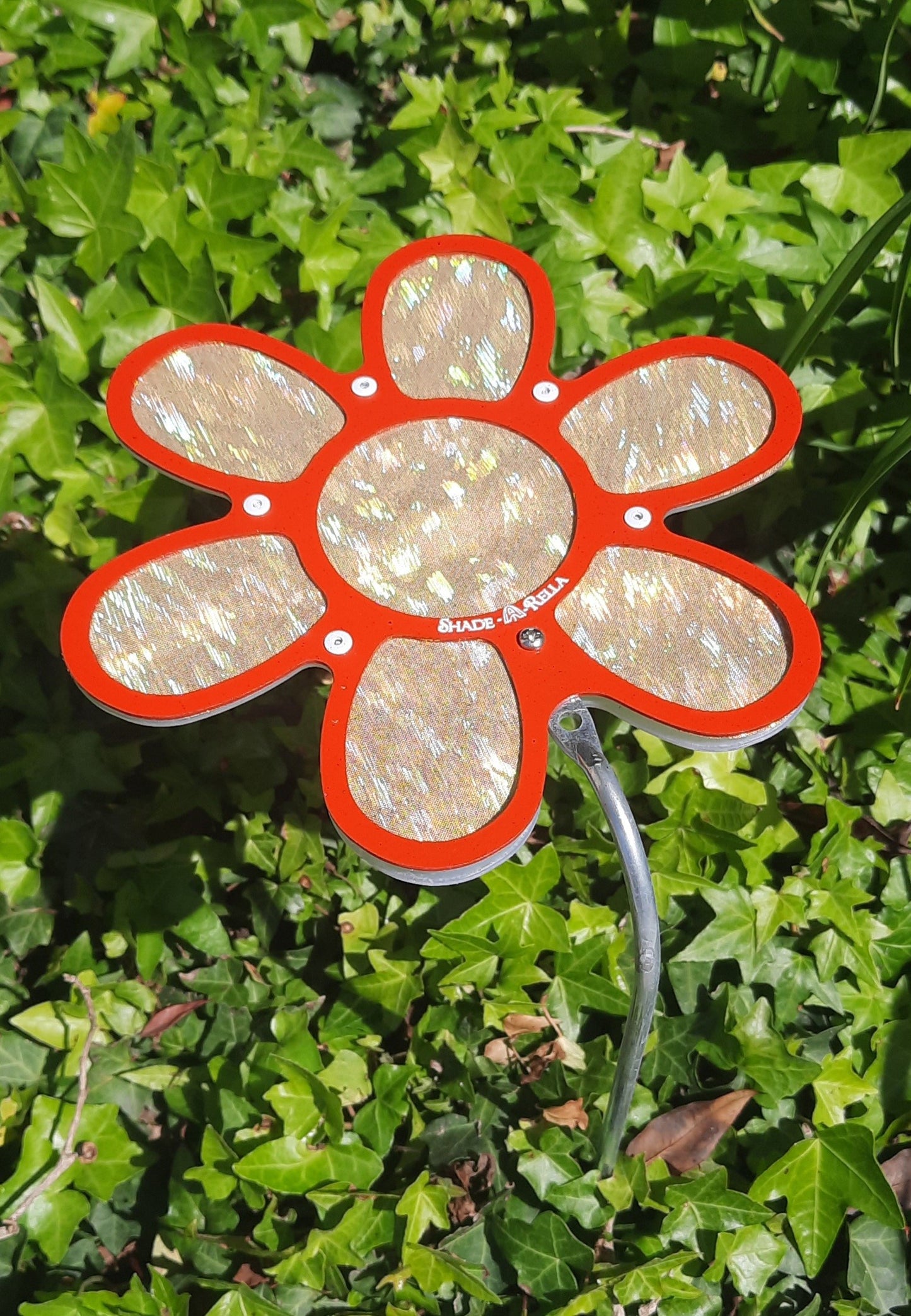 The 7.5 small Shimmer Flowers plant shade by Shade-A-Rella features iridescent cloth petals with a red outline and stands among green ivy, serving as both a decorative garden ornament and plant umbrella.