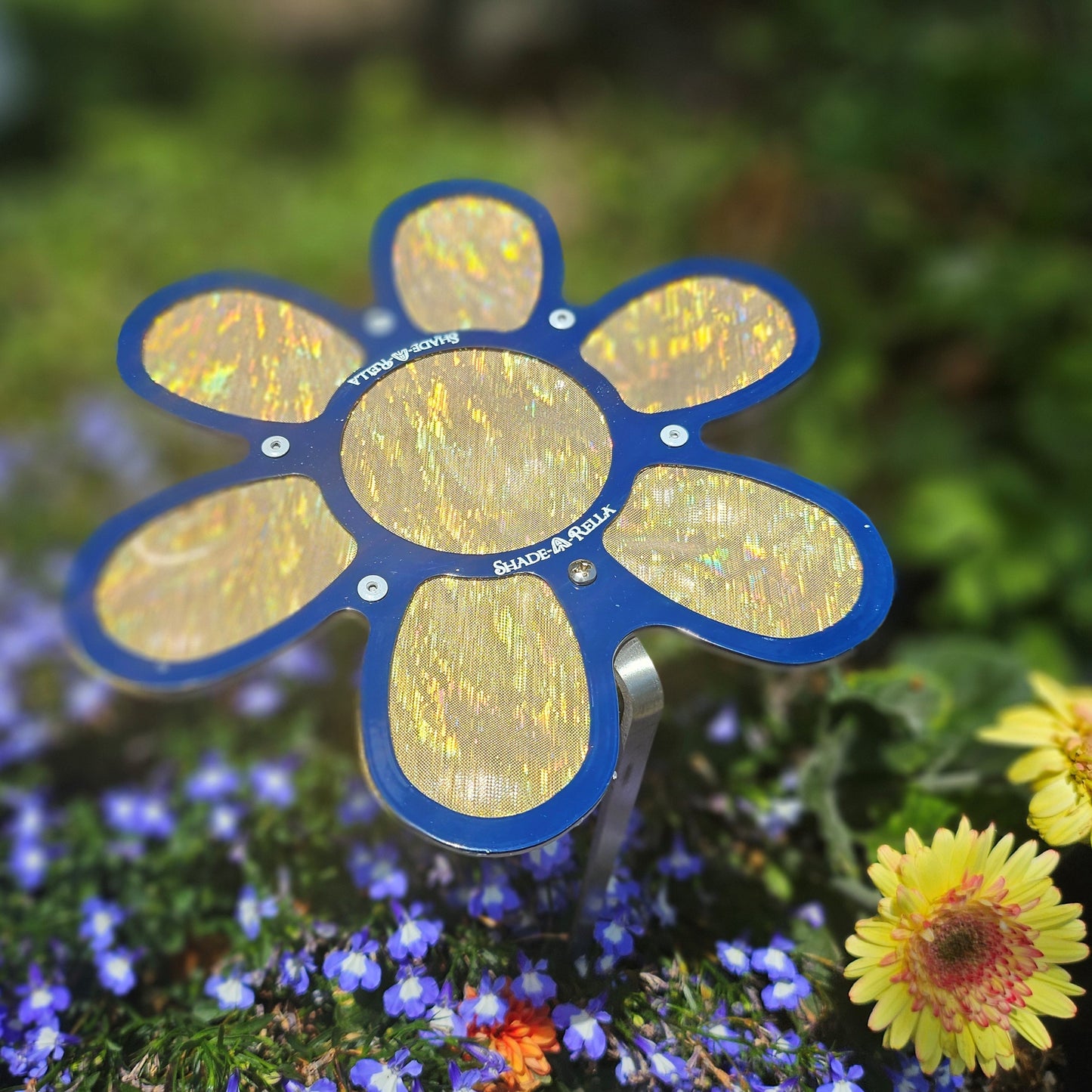 The Shade-A-Rella 7.5 small Shimmer Flowers plant umbrella features reflective yellow petals with a blue outline, sitting among blooms to offer plant shade. Its shimmering surfaces display text labels in your garden.