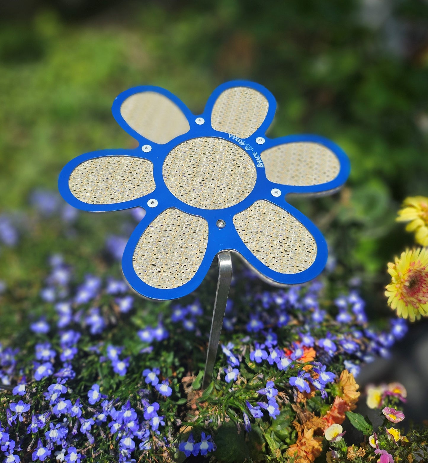 A 7.5 inch Shade-A-Rella Flower, blue with yellow/tan 86% shade cloth, is staked among blooming purple and yellow garden flowers with a blurred green foliage background.