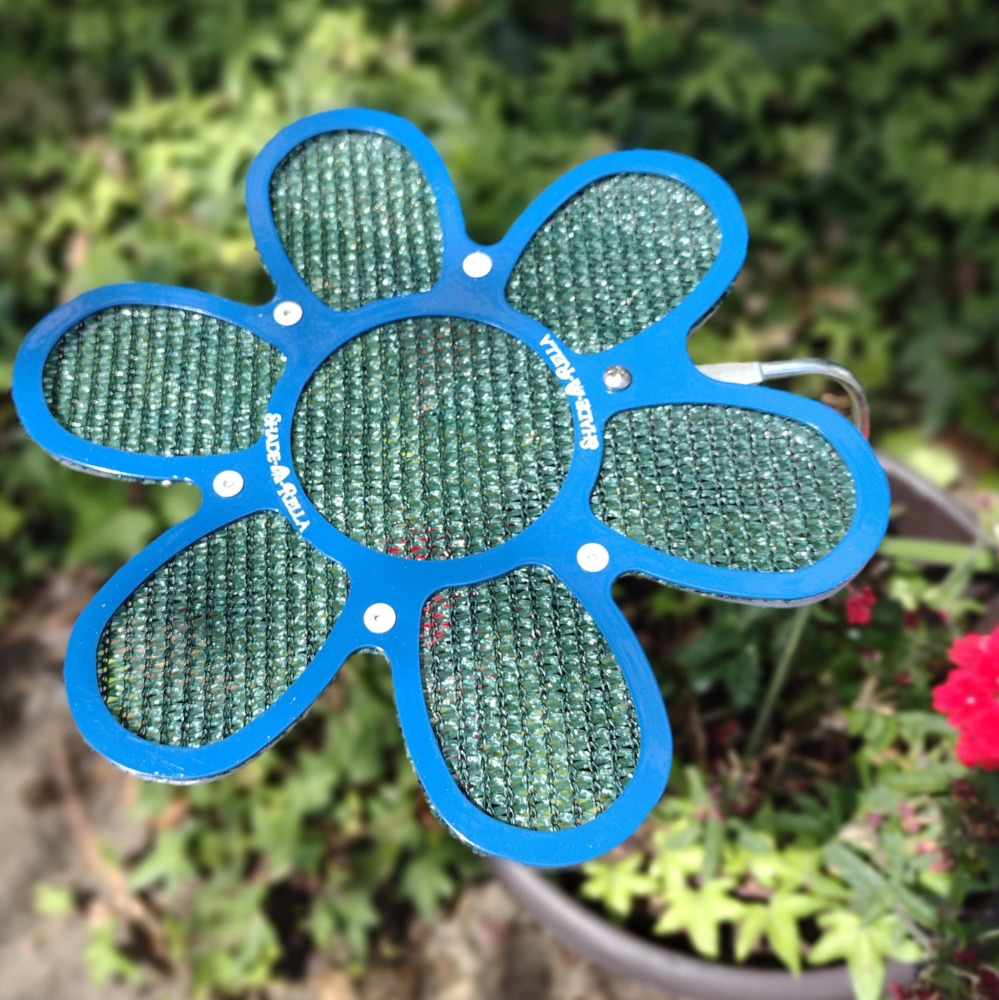 A 7.5 inch small blue flower-shaped mesh by Shade-A-Rella, with a blue frame and green 86% shade cloth, stands on a metal stem in the garden, creating an appealing plant shade near the window box amid greenery and red flowers.