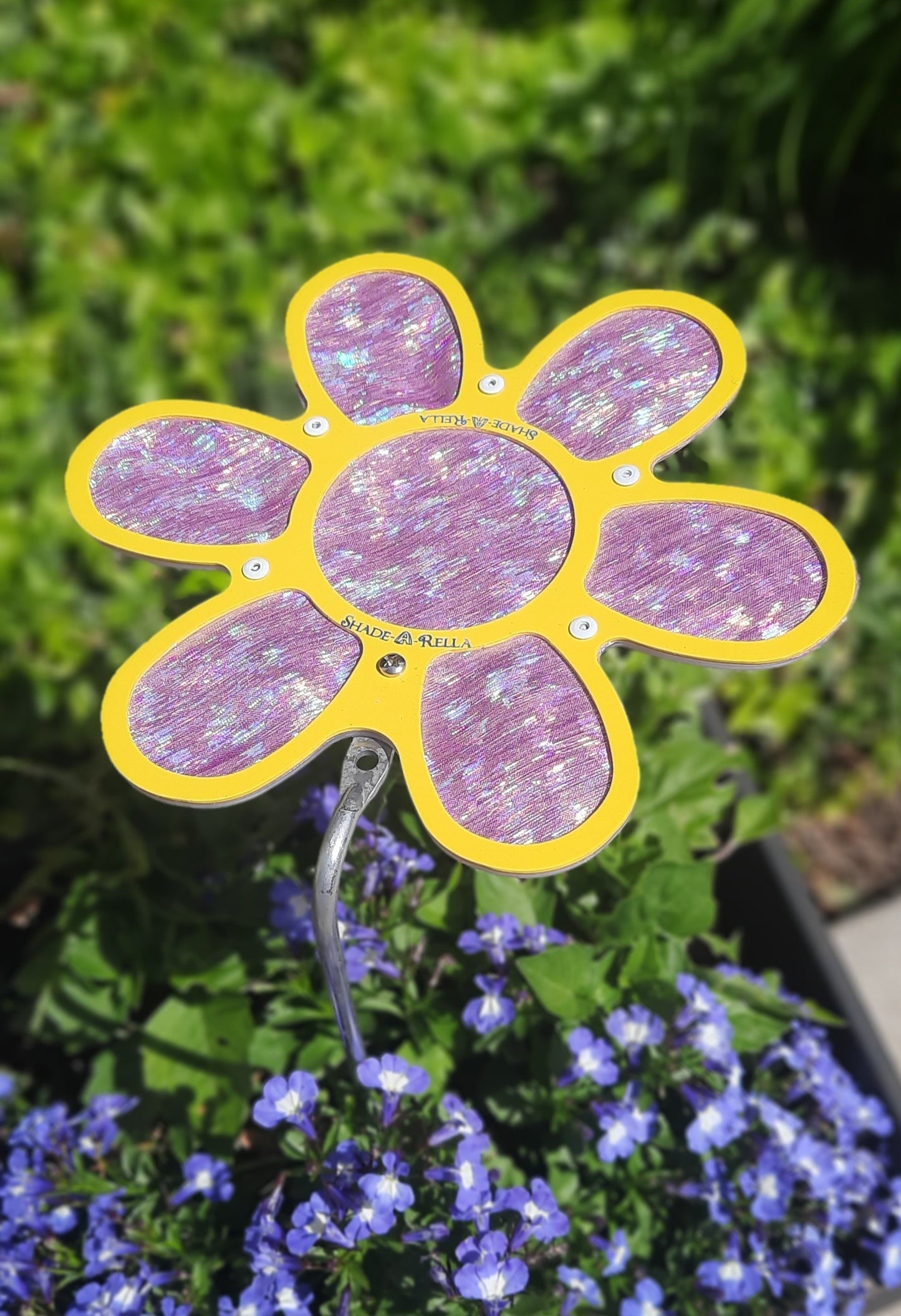 A 7.5 small Shimmer Flowers plant umbrella by Shade-A-Rella, with glittery purple petals and a hint of shimmer cloth, is placed among green foliage and small purple flowers in a garden.