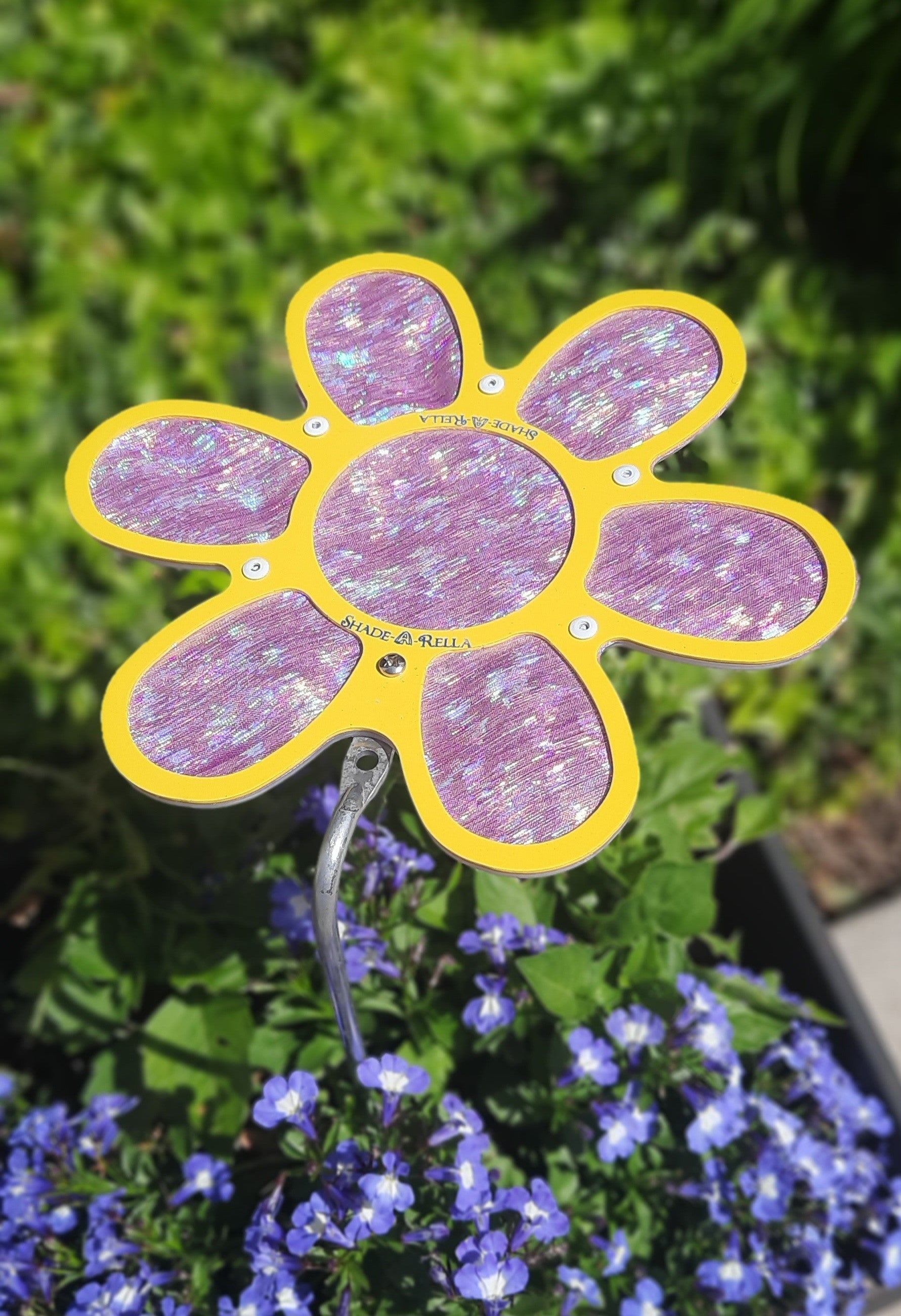 A 7.5 small Shimmer Flowers plant umbrella by Shade-A-Rella, with glittery purple petals and a hint of shimmer cloth, is placed among green foliage and small purple flowers in a garden.
