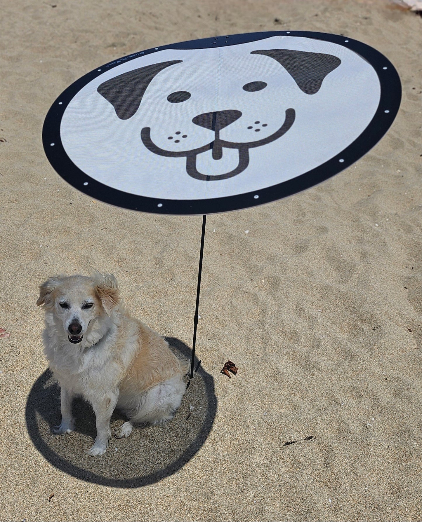 A fluffy, light-colored dog sits under the Shade-A-Rella Pup-A-Rella—a limited edition, 24" round portable shade with a printed puppy face on 70% shade cloth—casting a perfect shadow that frames the real pup beneath.