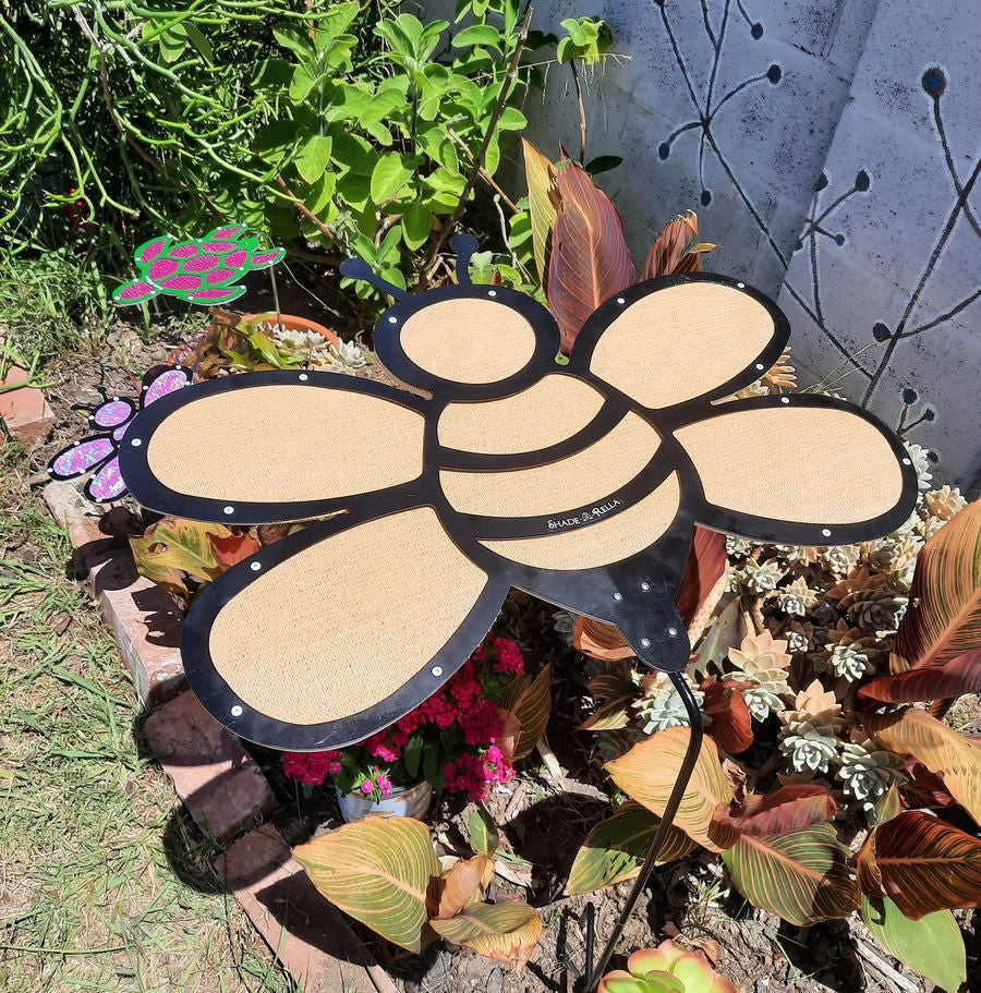 The Shade-A-Rella 24-inch Bee with black frame and light yellow 90% shade cloth stands among leafy plants and succulents, while a pink and green turtle ornament appears in the background against a white wall.