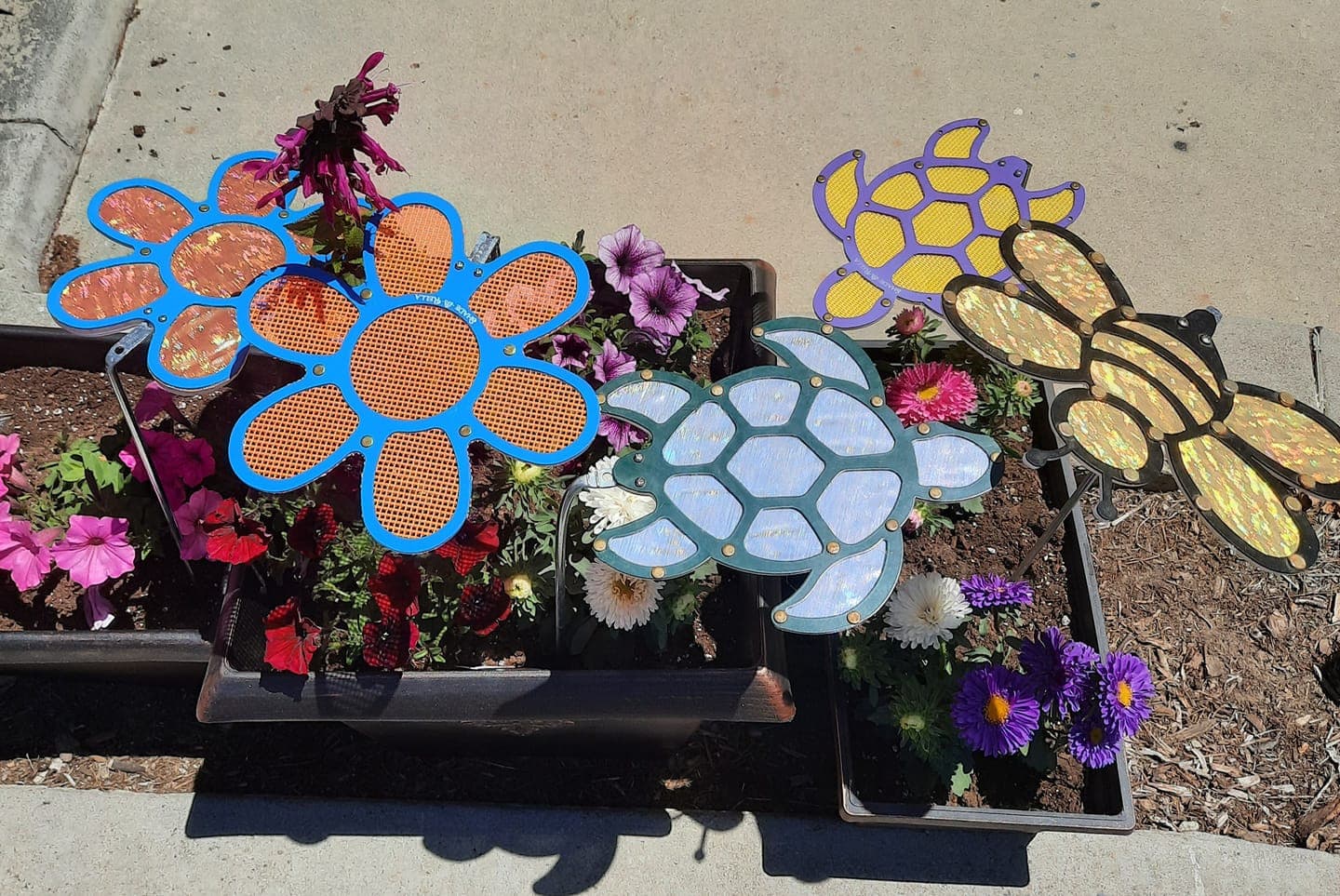 Shade-A-Rella’s 7.5 inch small Flower, featuring a red frame with blue 86% shade cloth, adds whimsical shade in planters on a sunny sidewalk, brightening the display among colorful flowers and decorations.