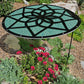 The Shade-A-Rella Green 24" Mandala plant shade—crafted with 90% shading cloth—stands among red daisies and lush foliage in a mulched garden bed, providing stylish protection for plants. Rocks and other greenery appear in the background.