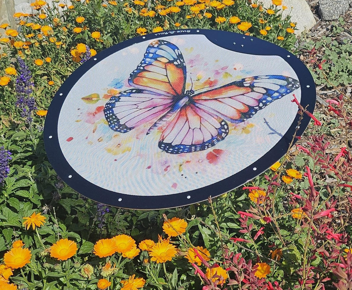The 24" Butterfly Shade-A-Rella by Shade-A-Rella is a durable plant umbrella featuring a colorful butterfly design and 70% shading cloth, providing garden shade among blooming yellow, orange, and red flowers.