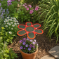 A garden scene features a Shade-A-Rella 7.5" small flower (red) on an 18" stainless rod in a pot with mulch, beneath green 90% shading cloth, surrounded by blooming flowers, greenery, and a stone pathway.