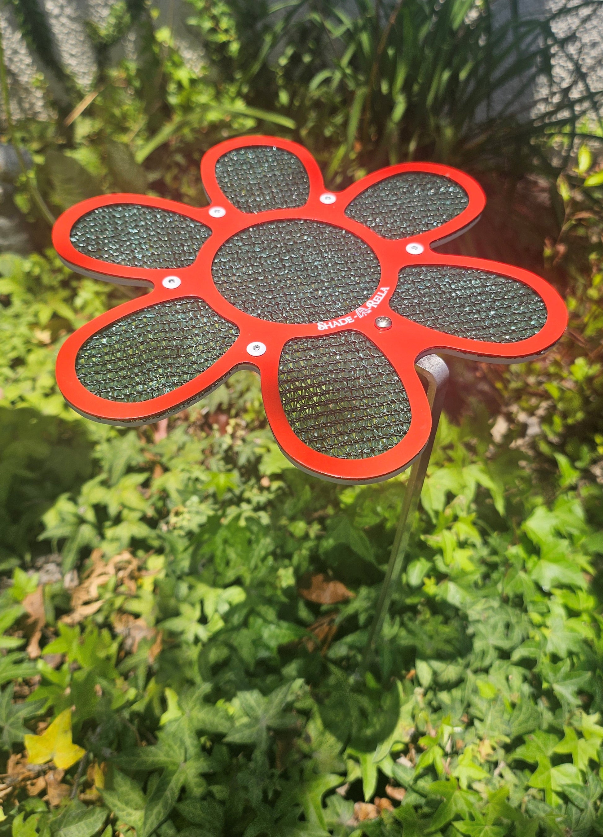 A 7.5" red Shade-A-Rella small flower with green 90% shading cloth and an 18" stainless rod stands in a garden bed, surrounded by green leafy plants and sunlight.