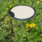 The Circle Shade-A-Rella by Shade-A-Rella is an adjustable plant umbrella with a beige top and black edge, featuring a powder-coated frame. Outdoors, it stands among green plants and yellow flowers with the SHADE-A-RELLA label on its edge.