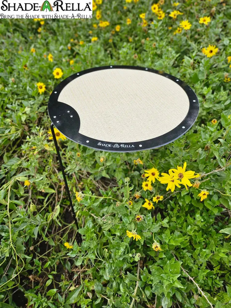 The Circle Shade-A-Rella by Shade-A-Rella is an adjustable plant umbrella with a beige top and black edge, featuring a powder-coated frame. Outdoors, it stands among green plants and yellow flowers with the SHADE-A-RELLA label on its edge.