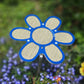 A 7.5-inch blue flower-shaped plant shade by Shade-A-Rella, made with yellow/tan 86% shade cloth, is mounted on a metal stake in a garden surrounded by green plants, window boxes, and small purple flowers.