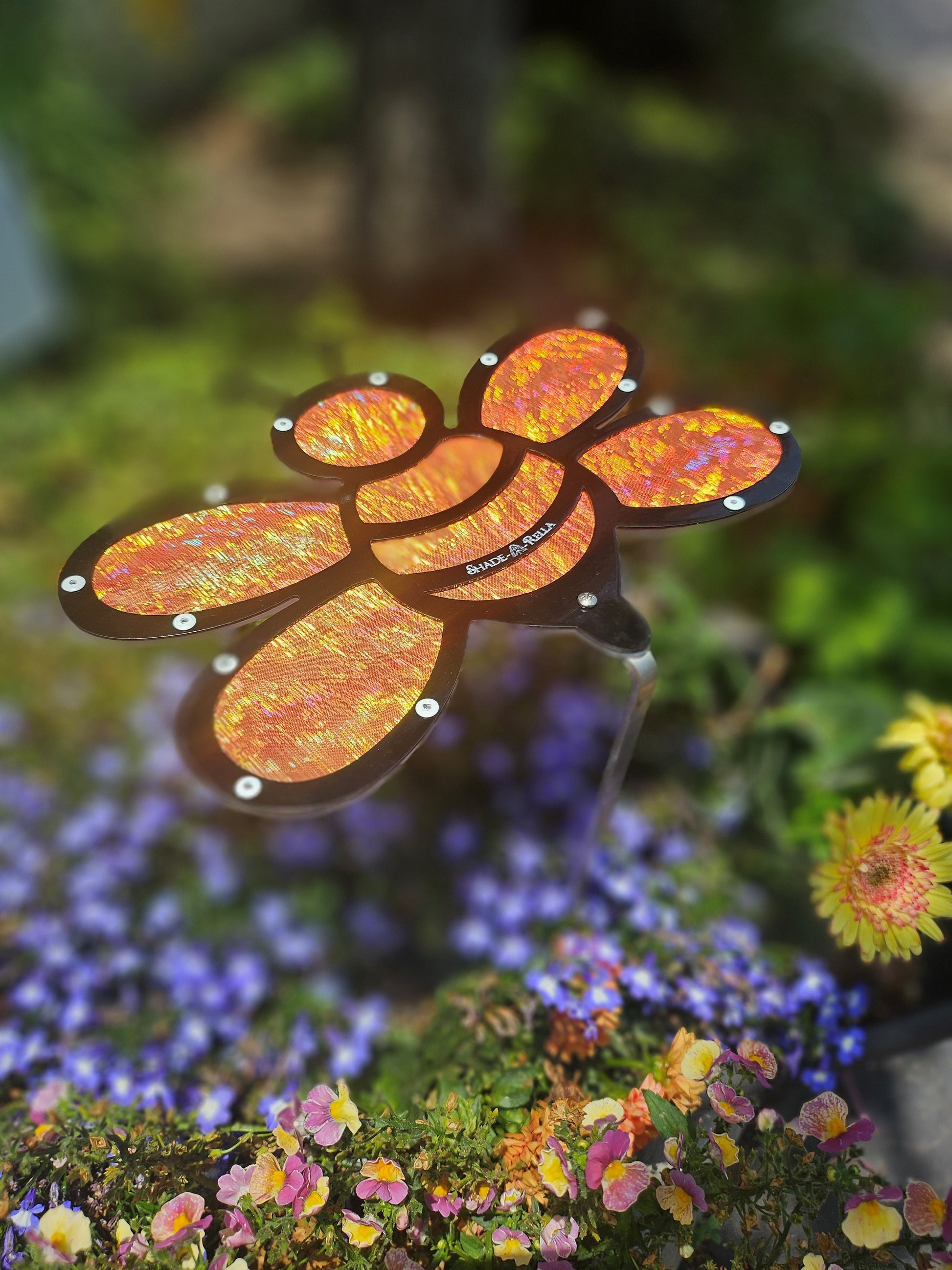 The Shade-A-Rella 10 inch small Bee with Orange shimmer cloth serves as a 100% shade cloth holder and Halloween bee, adding a metallic touch among colorful garden flowers and greenery.