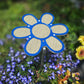 A 7.5 inch Shade-A-Rella Flower, blue with yellow/tan 86% shade cloth, is staked among blooming purple and yellow garden flowers with a blurred green foliage background.