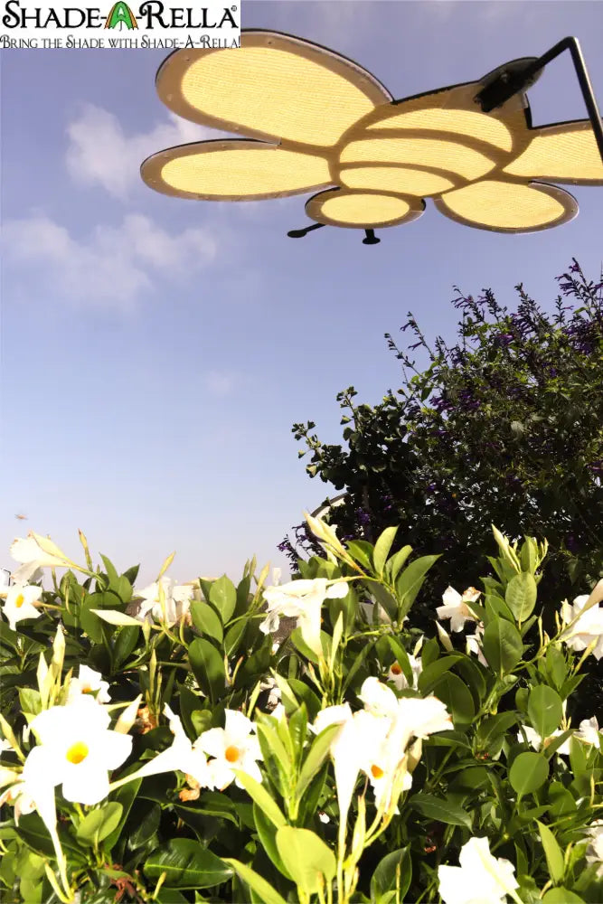 A Shade-A-Rella Super Sale! 24 inch Bee with black frame and light yellow 90% shade cloth hangs above lush bushes with white blooms, beside a subtle shade cloth holder, under a blue sky dotted with clouds.