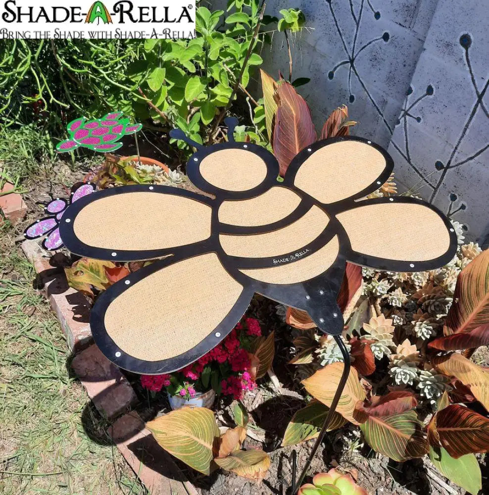 The Shade-A-Rella 24-inch Bee with black frame and light yellow 90% shade cloth stands among leafy plants and succulents, while a pink and green turtle ornament appears in the background against a white wall.