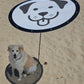 A fluffy, light-colored dog sits under the Shade-A-Rella Pup-A-Rella—a limited edition, 24" round portable shade with a printed puppy face on 70% shade cloth—casting a perfect shadow that frames the real pup beneath.